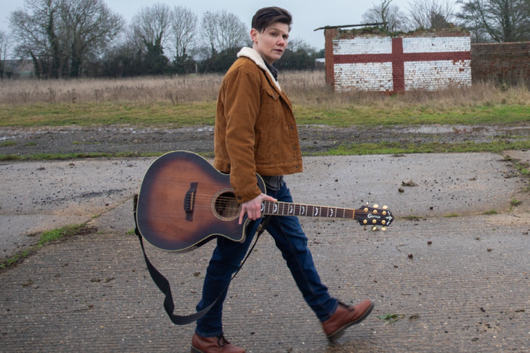 Folk singer Grace Petrie walking outdoors in a brown jacket, carrying an acoustic guitar by her side in a rural setting.